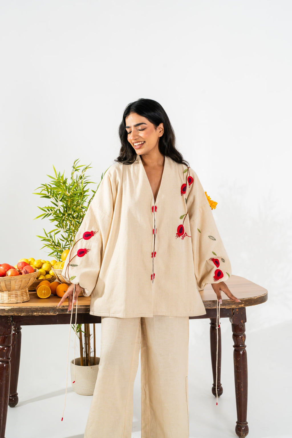 Woman in a beige outfit with red floral embroidery sitting at a table with fruits and plants.